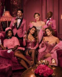 Diverse cabaret cast in pink costumes posed around velvet seating in a glamorous lounge