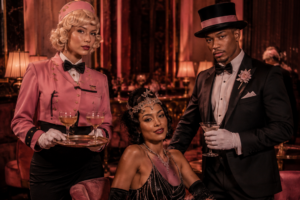 Cocktail server seated showgirl and tuxedoed host in a glamorous pink lounge